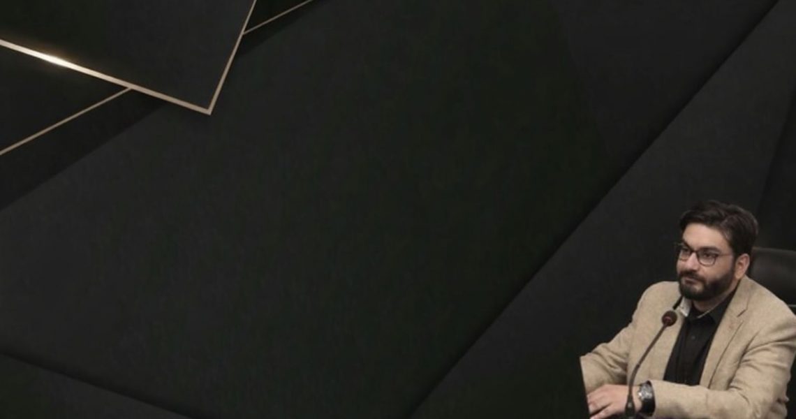 Ein Mann sitzt an einem Tisch vor einem dunklen, geometrischen Hintergrund und spricht in ein Mikrofon.