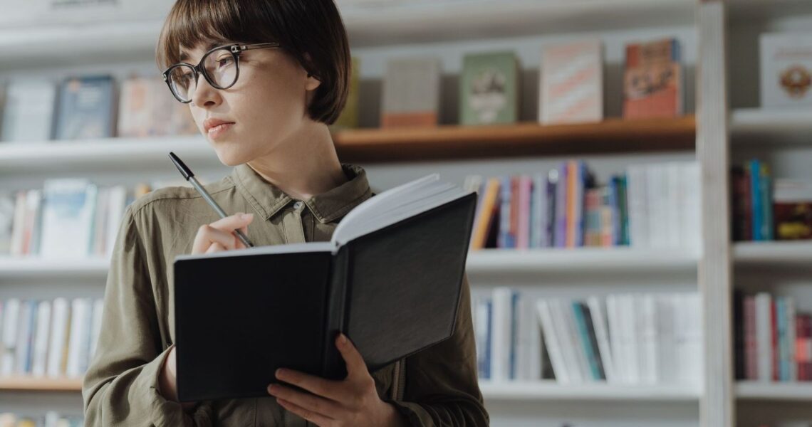 Frau mit Brille und Notizbuch vor einem Regal voller Bücher